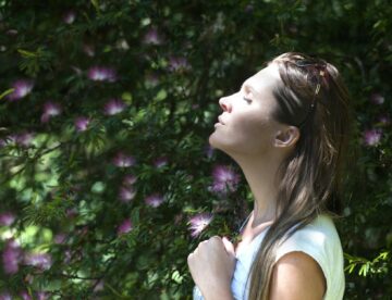woman closing her eyes against sun light standing near purple petaled flower plant